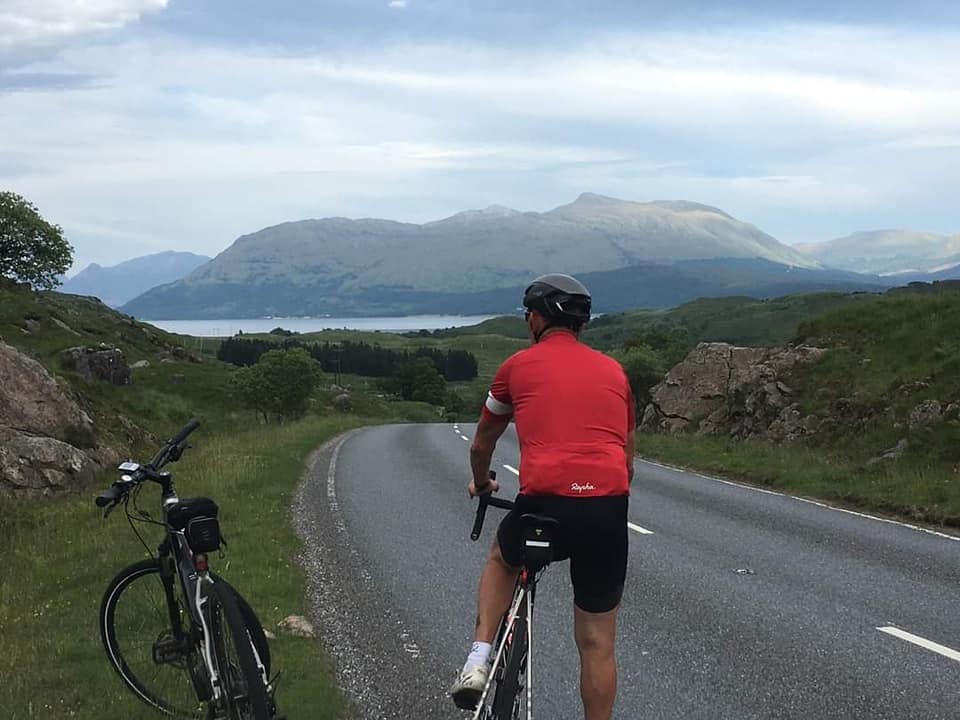A member of 605 Squadron cycles along a road with scenic hills and landscapes ahead of him