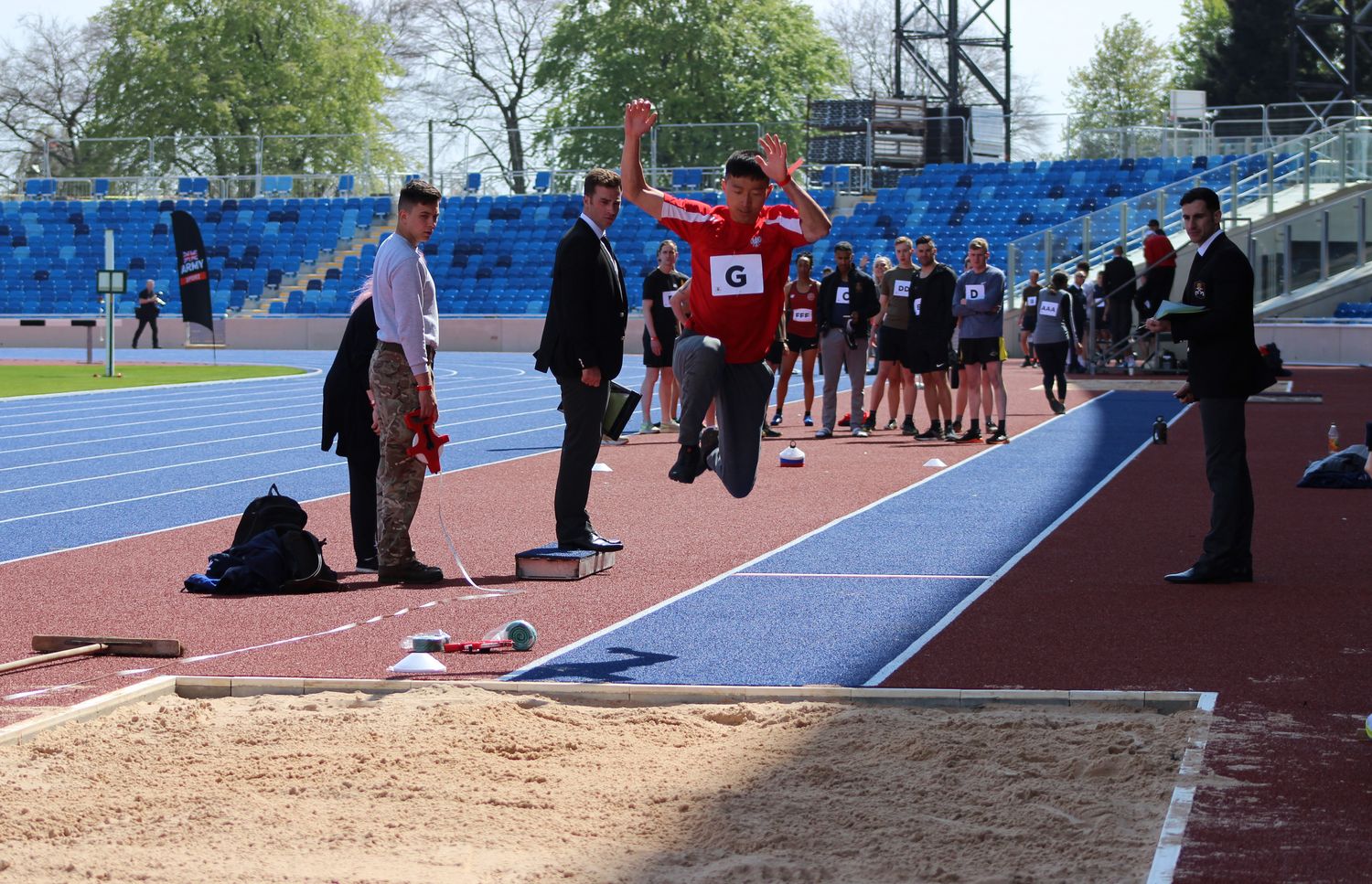 An Army athlete is mid-jump through the long jump at the Midlands Army Athletics Championships at the Alexander Stadium, watched by judges and other competing members from the Army