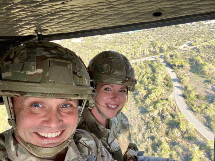 Two Reservists in combat uniform are smiling towards the camera as they take a selfie whilst flying in the air from a helicopter, with trees and landscapes visible from the helicopter.