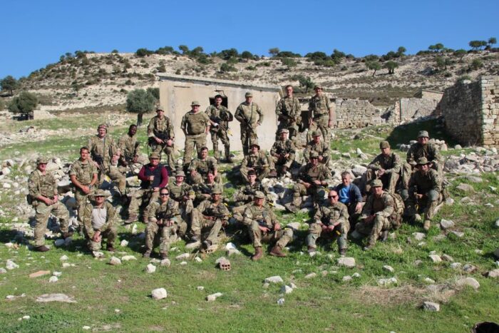 Reservists from 37th Signal Regiment are gathered together in their uniforms after completing a section attack for Exercise Kronos Hunter. They are sitting and standing on rocks on the ground in front of a ruined stone building.