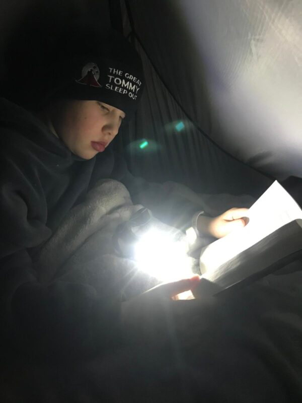 Cadet Moody sits in his tent, wrapped up in a hat and blanket. He is reading a book in the dark with the help of a torch.