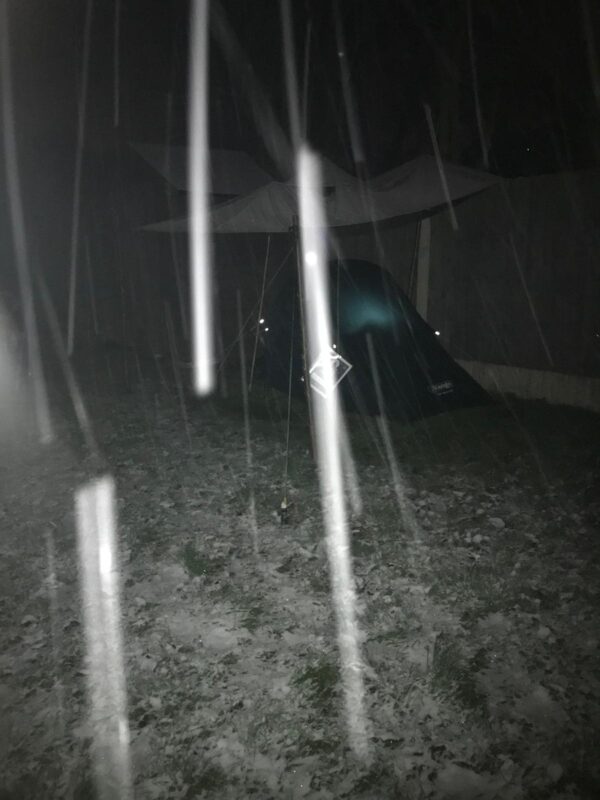 The tent is pitched in a garden, in the dark. Snow is falling from the sky and settling on the tent and grass around it.