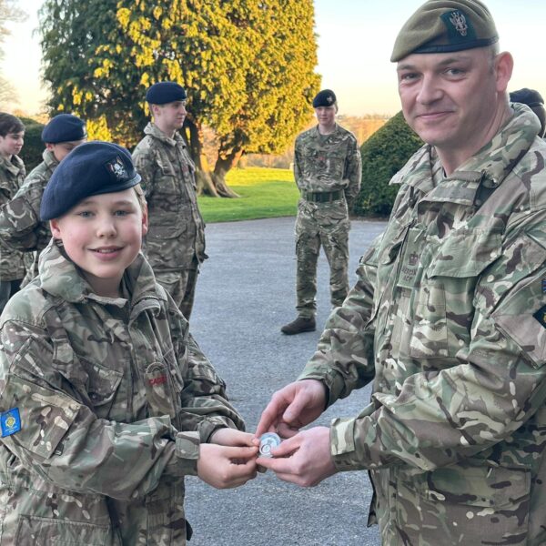 Cadet Moody in his Army Cadet Force uniform, smiling as he is presented with a special embossed metal coin by their Officer Commanding. His fellow cadets stand in the background in the evening sunshine, watching the presentation.