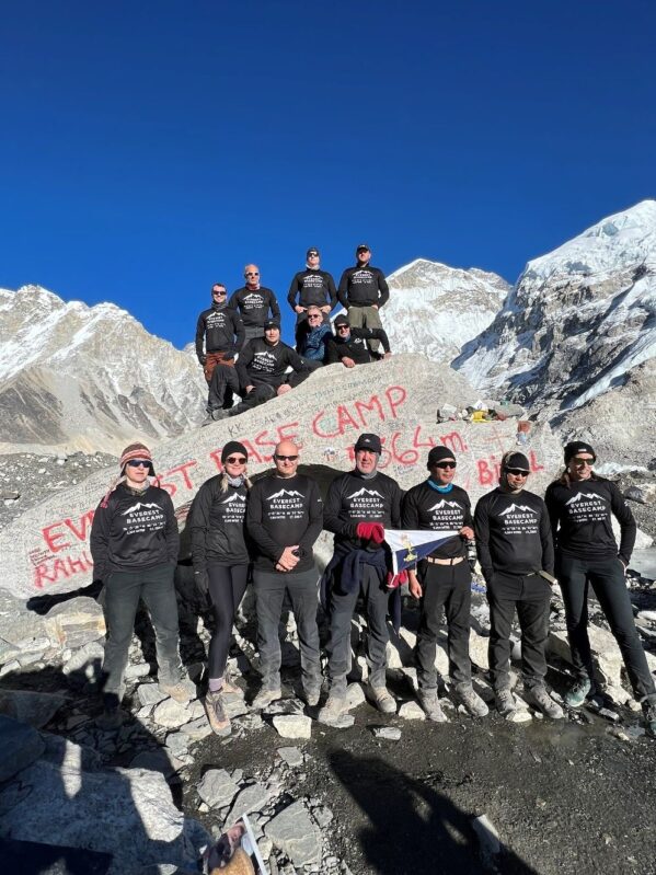 Fourteen Reservists made it to the Everest Base Camp. They are pictured smiling as a group, holding their Regimental flag, standing on rocks with blue skies in the background.