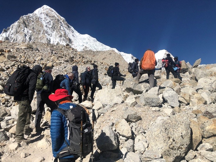 A group of Reservists in warm clothing and safety gear climb over rocks and rocky terrain as they make their ascent up towards Base Camp. The weather conditions are clear with sunshine and blue skies in the background.
