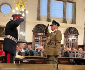 Captain Frith being presented his 4th Clasp for his service to the Army Cadets by the Lord-Lieutenant for Staffordshire. Both are in uniform, facing next each other and saluting, in front of guests sitting behind them.