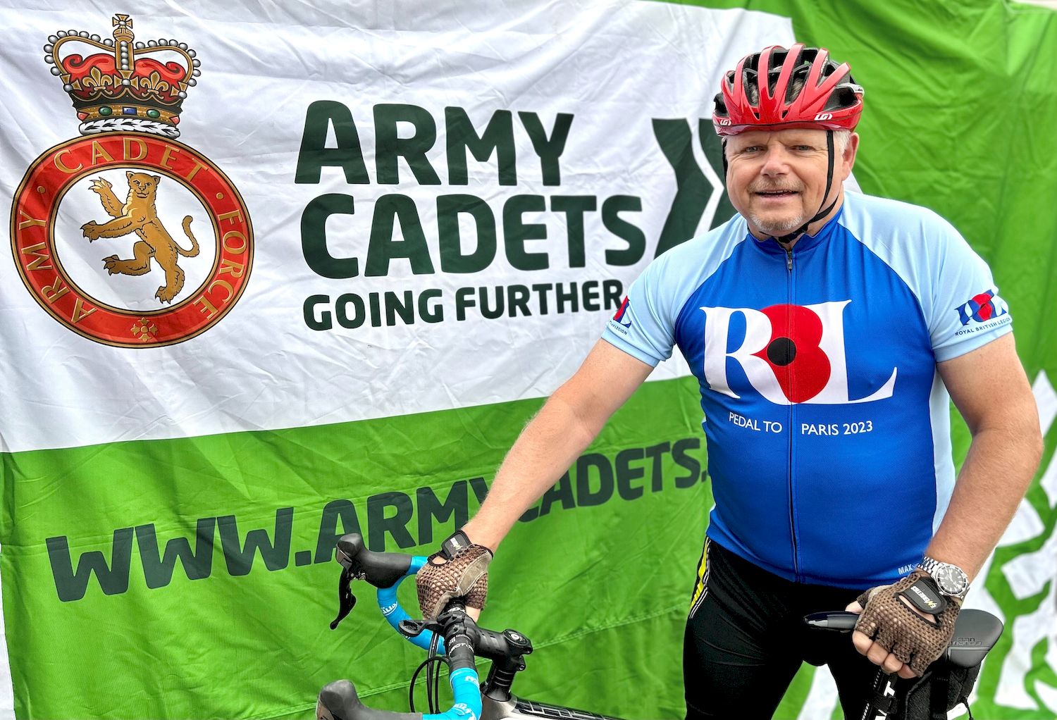 Jim wears his Royal British Legion cycling uniform, standing smiling with his bike in front of an Army Cadets banner