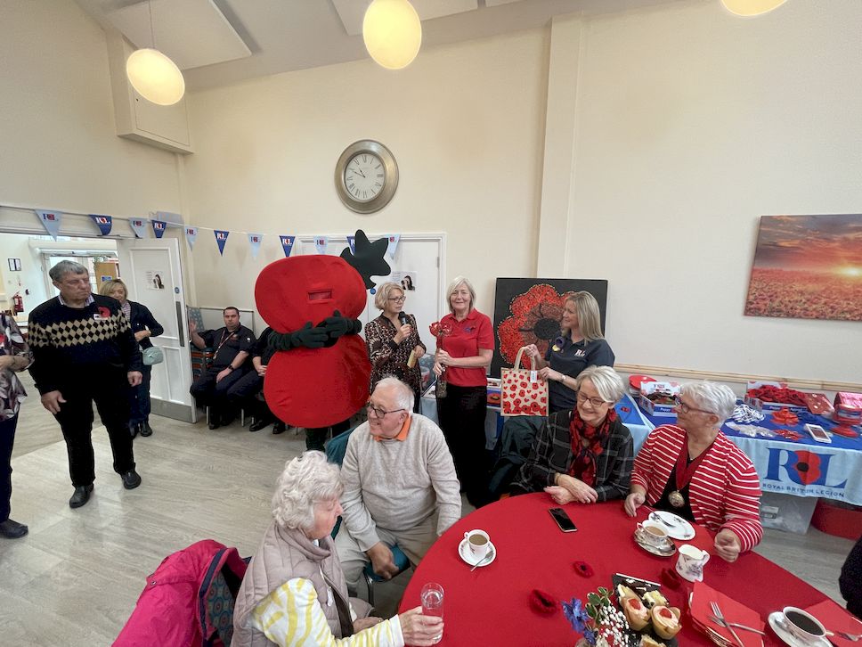 Guests at Galanos House are enjoying tea, live music and activities at Galanos House's Poppy Appeal launch. They are seated at tables smiling, enjoying activities, with the Poppy Appeal mascot in the background
