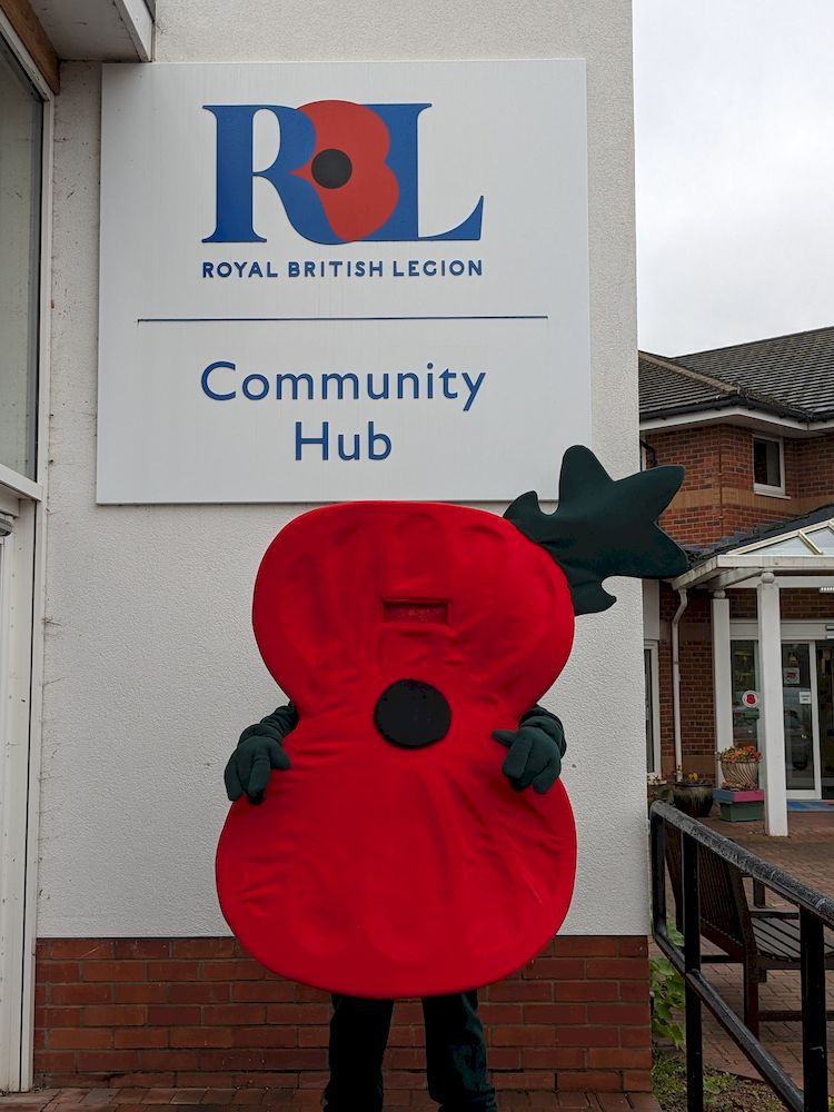 A mascot dressed up in a red poppy shaped outfit is standing outside the Galanos House Comunity Hub sign