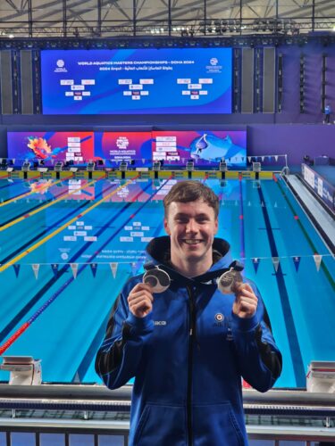 Air Specialist Class 1 Matt Holland from 605 Squadron is holding up two of his silver medals, smiling. Behind him is the indoor swimming pool in Doha which was used for the competition.