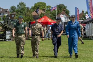 One member from each of the Royal Marines, British Army, Navy and Royal Air Force. They are walking across a field, with unit stand in the background) towards the camera.