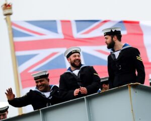 Three Naval personnel stood on ship with the Union Jack flag in the background.