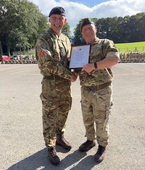 SMI Cartlidge in his Cadet Force Adult Volunteer uniform, receiving an award. he is shaking hands and smiling at the camera as he receives his certificate.