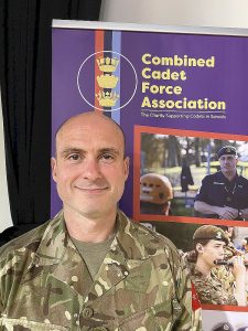 Corporal Mark Bailey in his Cadet Force Adult Volunteer uniform, standing in front of a banner advertising the Combined Cadet Force Association