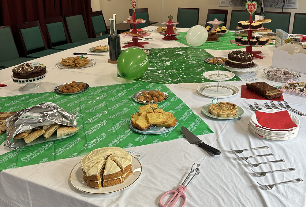 A table decorated with white and green cloths and balloons is laden with a huge variety of cakes and savoury items donated by our team members, ready to raise funds for Macmillan Cancer Support